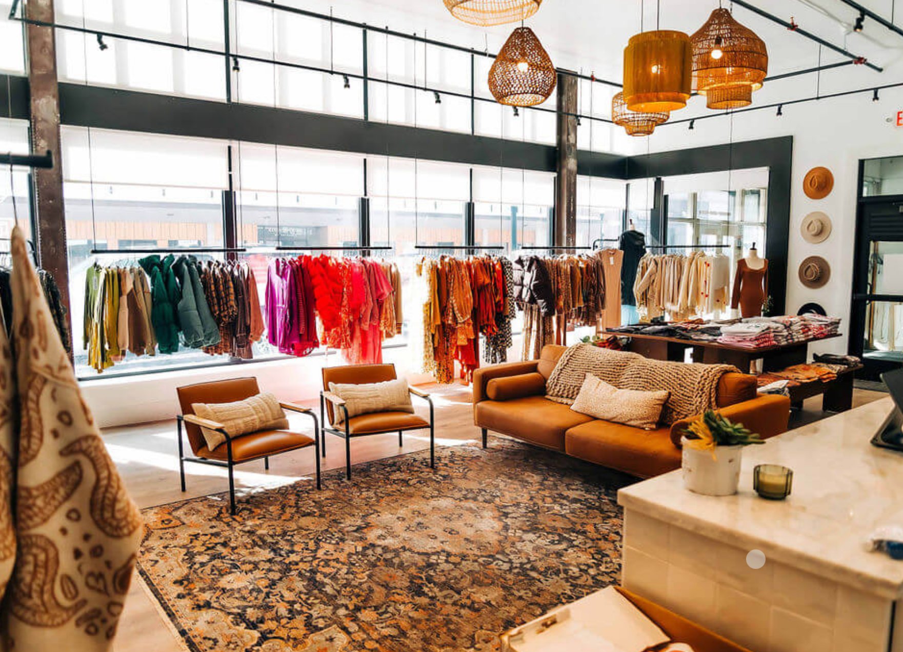 Modern boutique interior with clothing racks, tan leather seating, pendant lighting, and floor-to-ceiling windows overlooking a city skyline
