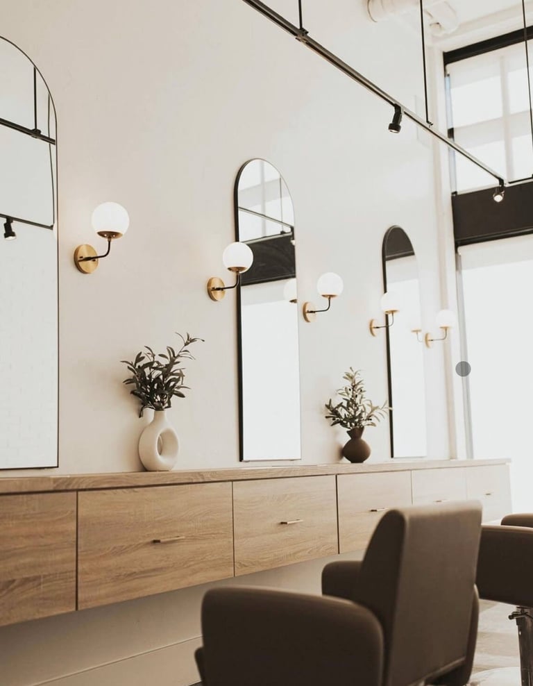 Modern salon interior with wooden vanity, arched mirrors, brass sconces, potted plants, and a dark upholstered chair