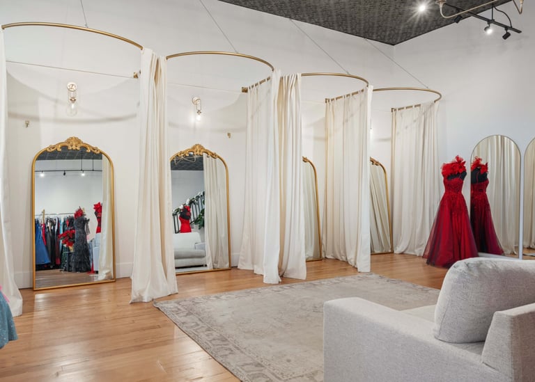 High-end retail dressing room with multiple fitting stalls separated by cream curtains, wooden frames with mirrors, formal red gowns displayed, wooden flooring, and upholstered seating
