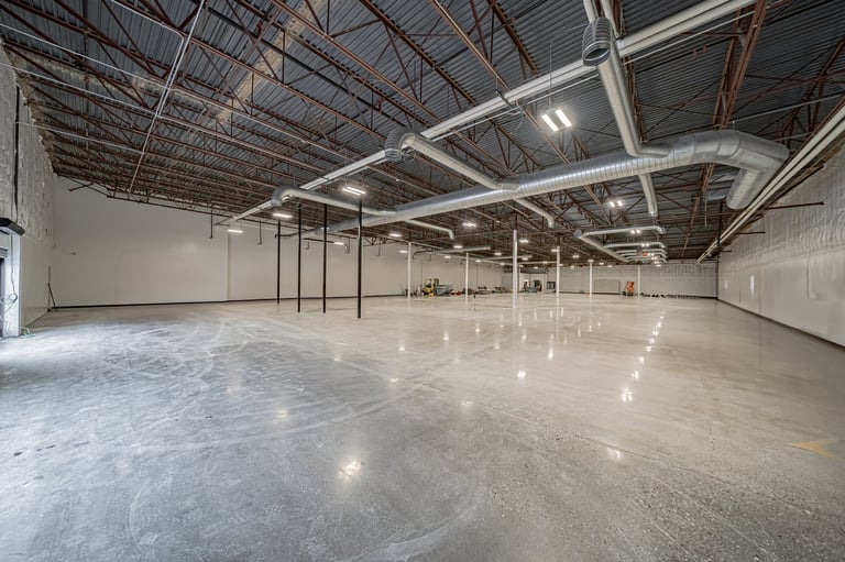 Empty industrial warehouse interior with polished concrete floor, exposed ceiling beams, metal pillars, and bright LED lighting