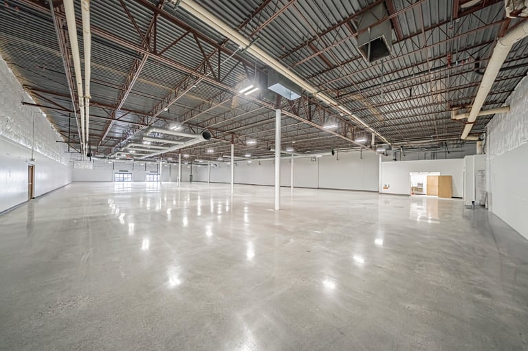 Empty industrial warehouse interior with polished concrete floor, white walls, metal ceiling beams, and overhead lighting