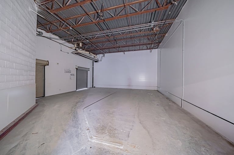 Empty industrial warehouse space with concrete floor, white walls, exposed metal ceiling beams, and doors on the left side