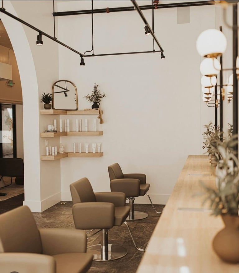 Modern salon interior with tan leather chairs, wooden shelves with products, industrial pendant lighting, and minimalist beige walls