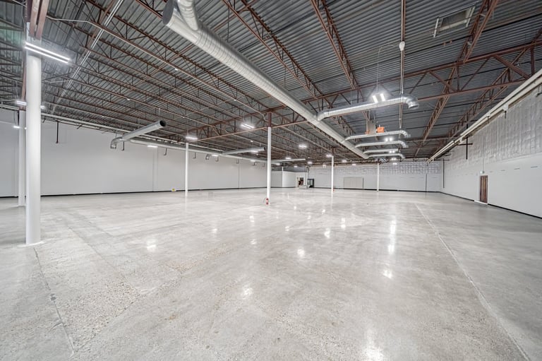 Empty warehouse interior with polished concrete floor, white walls, and metal beam ceiling structure with fluorescent lighting