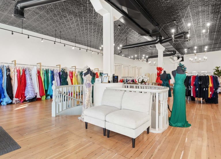 Modern boutique clothing store interior with colorful dresses on display racks, white seating, wooden flooring, and industrial ceiling