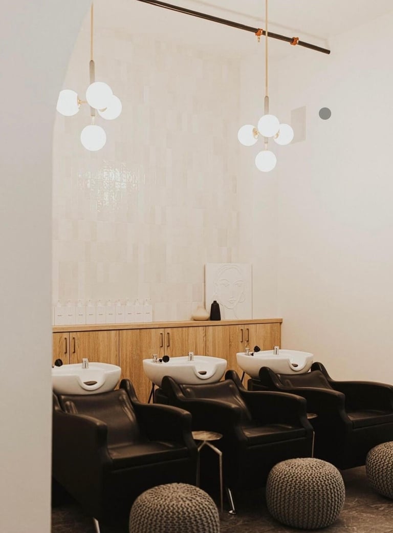 Modern salon interior with three black wash chairs, white sinks, wooden vanity, and hanging pendant lights with brass fixtures above