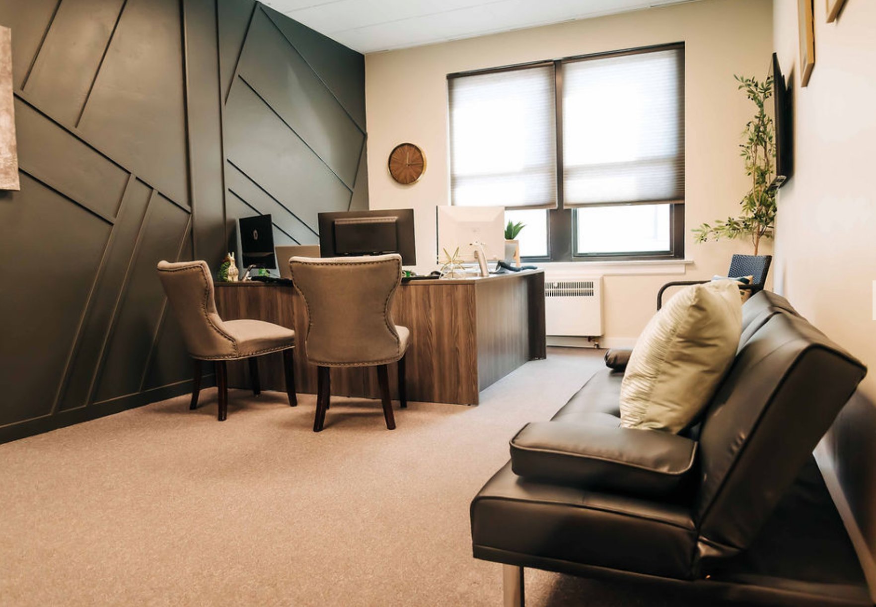 Modern office interior with black accent wall, two tan chairs at desk, and black sofa with cushions near windows